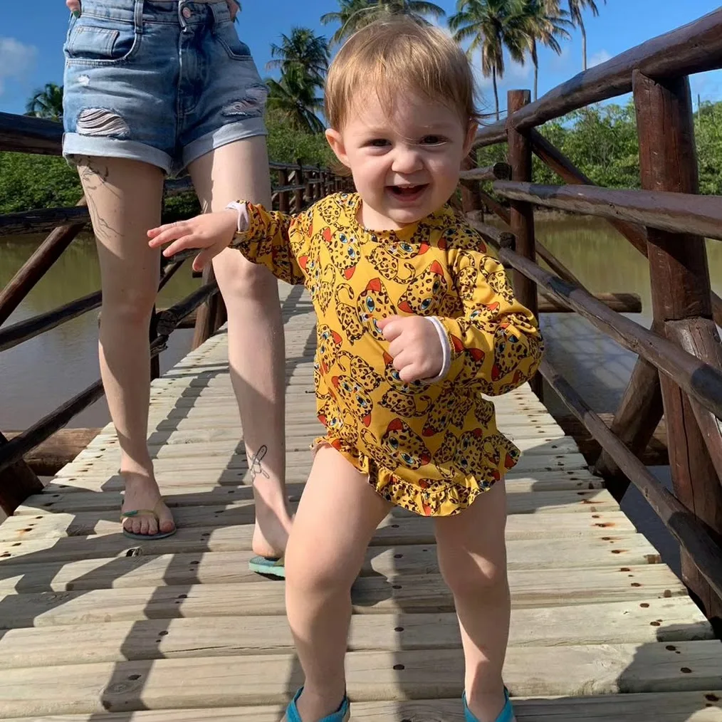 Menina pequena, sorrindo, caminhando de mãos dadas com um adulto numa passarela de madeira