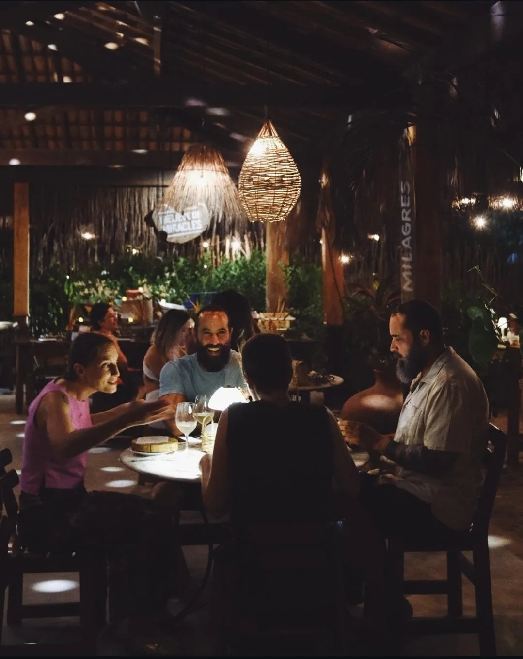 Friends at a dinner table at night under straw lamps, on the Route of Milagres
