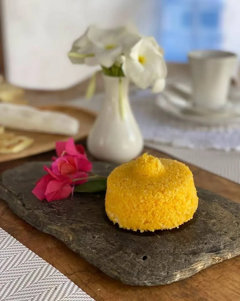 Café da manhã típico com cuscuz amarelo servido sobre uma pedra, com uma flor ao lado
