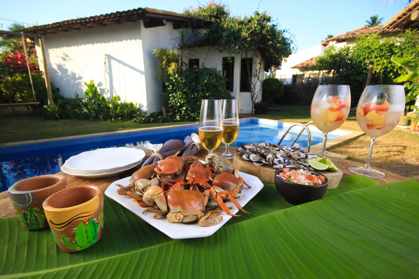 Mesa ao ar livre com petiscos sobre folhas de bananeira, ao lado da piscina do Pontal da Enseada